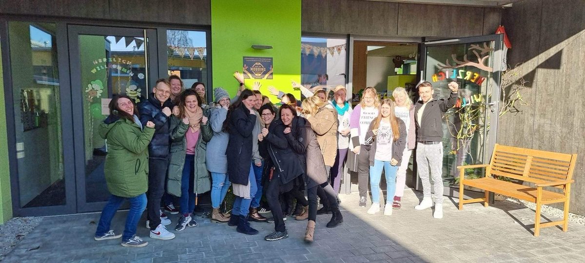 Das Team des Diakonie-Kindergartens Gräfenberg in Heldenpose vor der Einrichtung, an der Wand die EOM-Zertifikatstafel